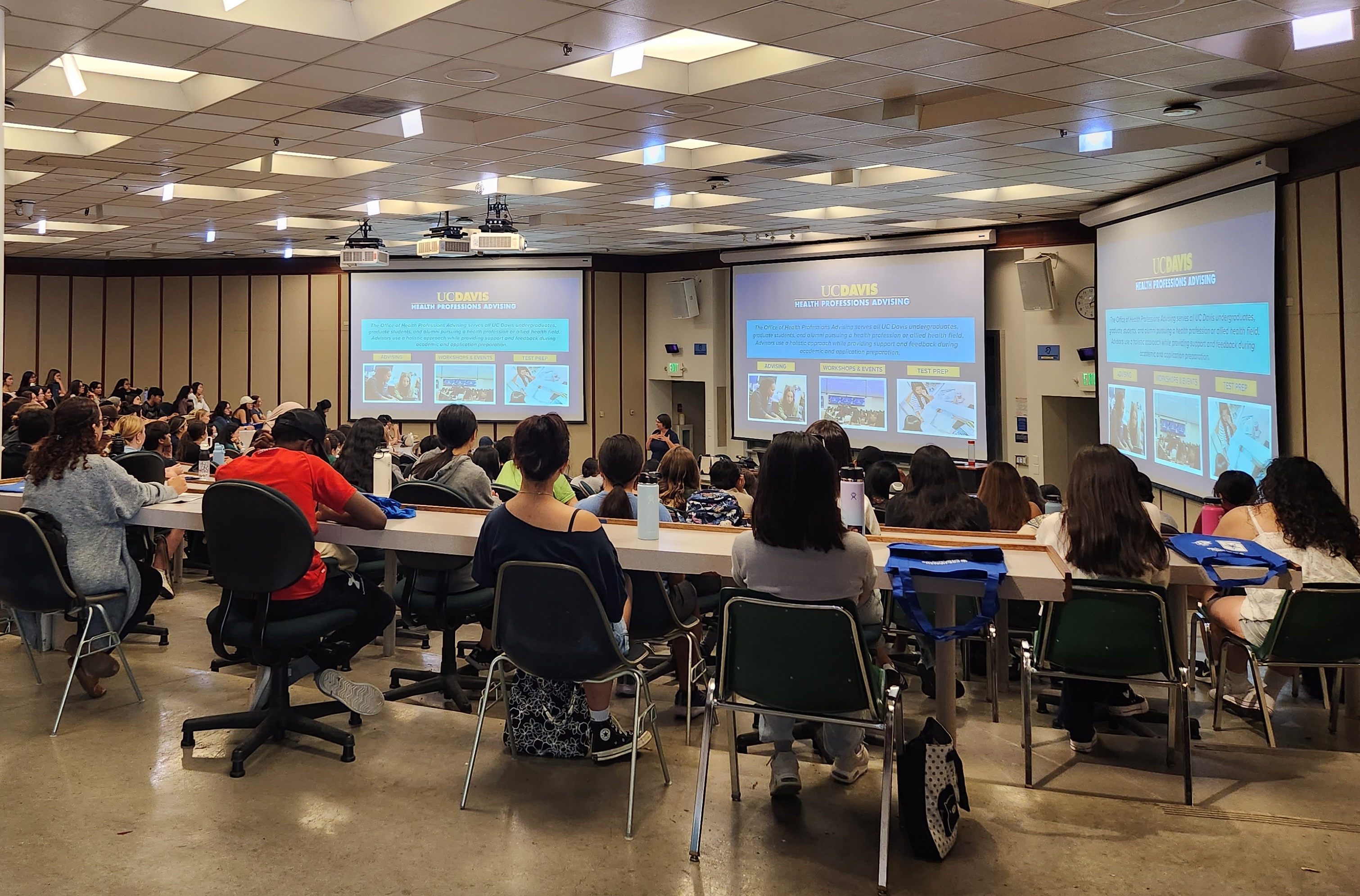 Many students sitting in a large lecture hall watching a presentation.