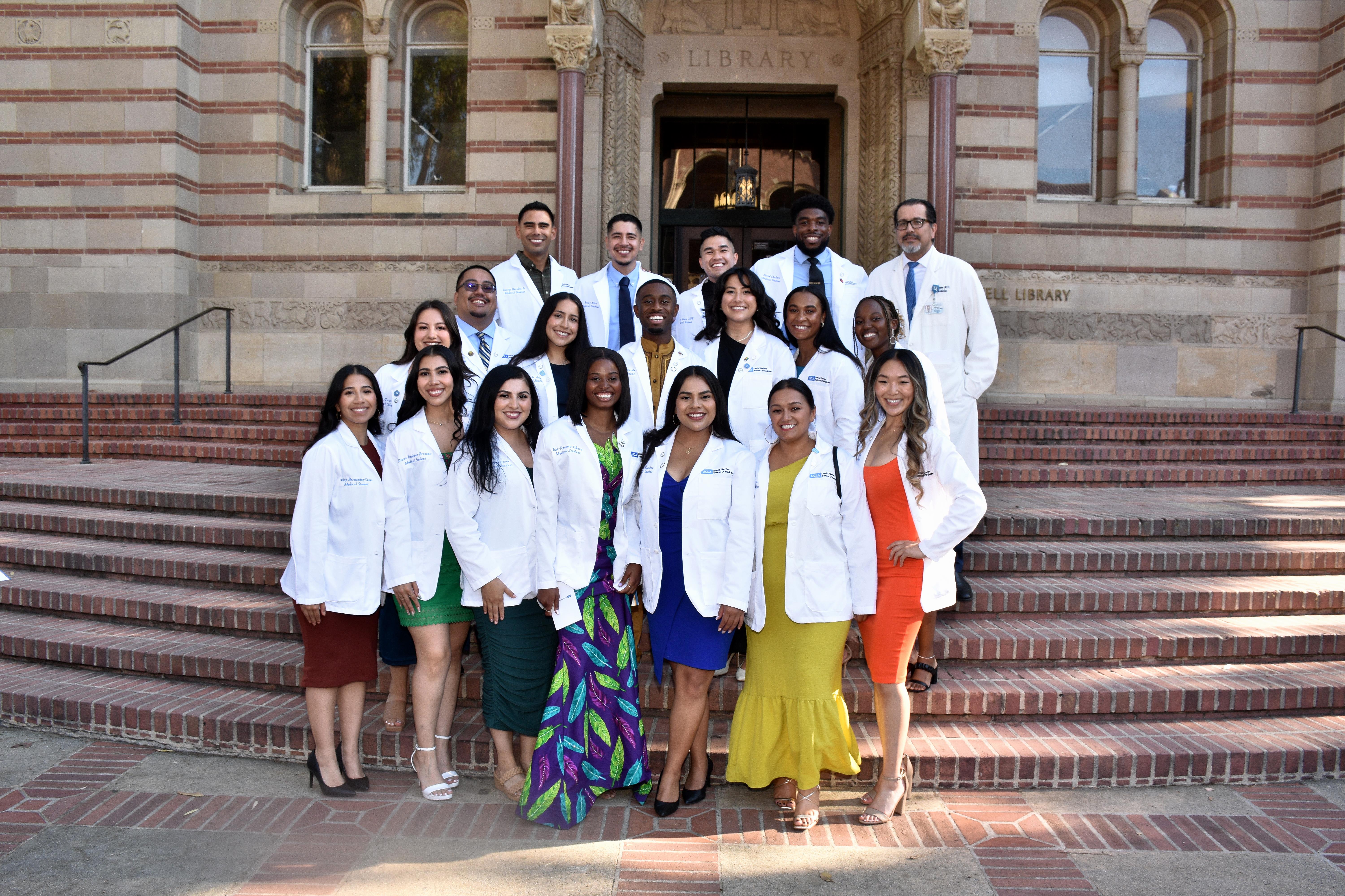 Group of medical students at PRIME-LA David Geffen School of Medicine gather on the steps in front of a building while wearing their white coats.