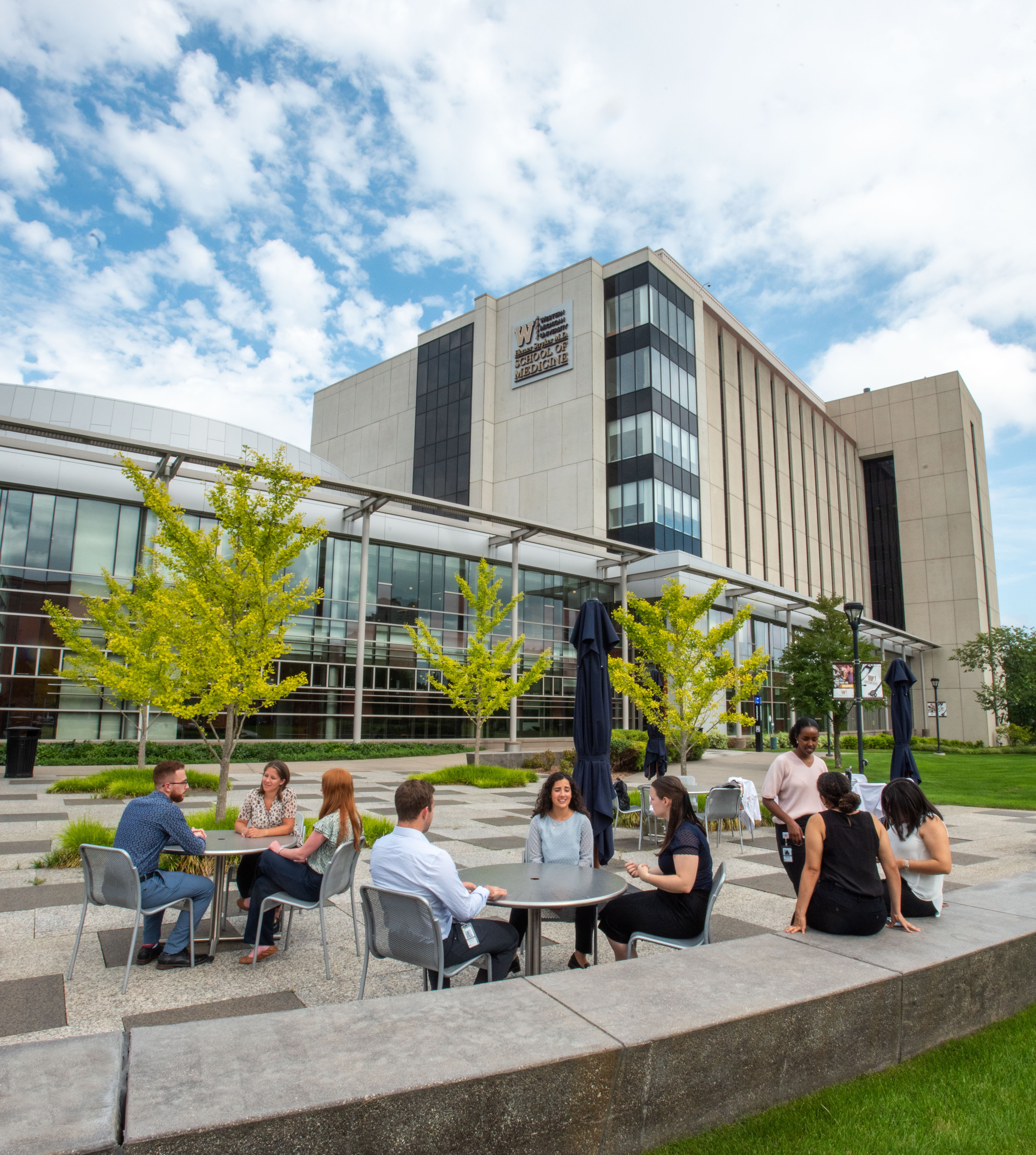 Groups of people gather around tables in front of a building on Western Michigan University Homer Stryker School of Medicine campus.