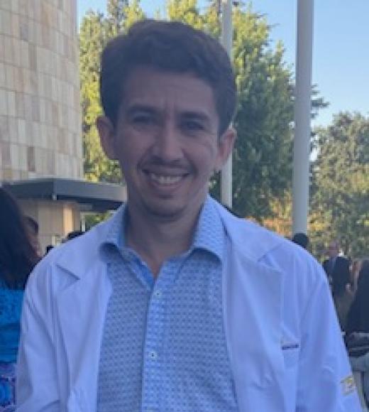 Image of pre-vet advisor Christian Guerzon. He is wearing a stripped button up shirt and lab coat. He is also wearing navy blue slacks with a brown belt and grey and white sneakers. He is standing near a building and in front of some trees and people.