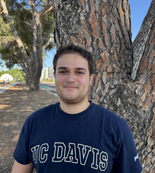 A photo of Christian smiling at the camera wearing a navy blue shirt in front of a tree.
