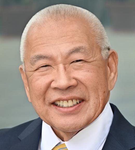 Image of Ronald S. Mito wearing a suit and blazer with a yellow, blue, and white striped tie. The right side of his blazer has a UCLA Dentistry Alumni pin and a smiling tooth pin. The background is blurry and grey.