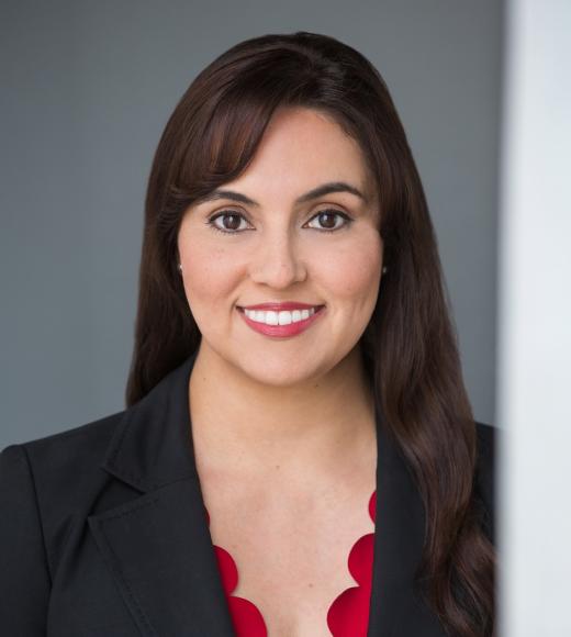 Image of Advisory Board member Monica Donnelley. She has on a red blouse and black blazer. She is standing in front of a grey wall and leaning on a white wall.