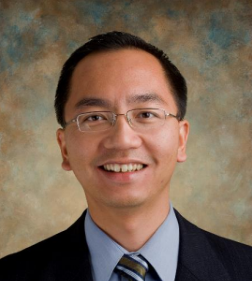 Image of John D. Vu in a blue dress shirt, tie, and black blazer. The background is a blur of brown and blue colors.