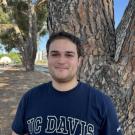 A photo of Christian smiling at the camera wearing a navy blue shirt in front of a tree.