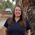 Smiling woman in navy polo standing by a large pine tree on a park path
