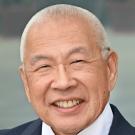 Image of Ronald S. Mito wearing a suit and blazer with a yellow, blue, and white striped tie. The right side of his blazer has a UCLA Dentistry Alumni pin and a smiling tooth pin. The background is blurry and grey.