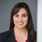 Image of Advisory Board member Monica Donnelley. She has on a red blouse and black blazer. She is standing in front of a grey wall and leaning on a white wall.