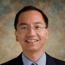 Image of John D. Vu in a blue dress shirt, tie, and black blazer. The background is a blur of brown and blue colors.